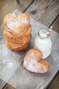A stack of icing sugar sprinkled, cherry heart-shaped donuts, with a milk bottle full of milk and an old fashioned paper straw.