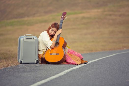Young hippie woman with guitar and backpack sitting on a countryside road