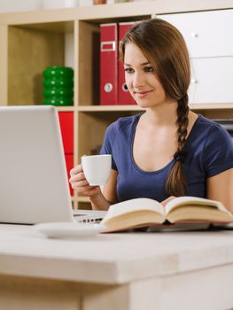 Photo of a beautiful smiling woman using a laptop and drinking coffee at home or at her office.