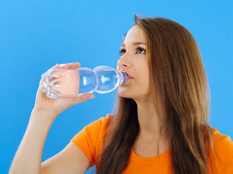 Photo of a beautiful young brunette woman drinking bottled water over blue background.