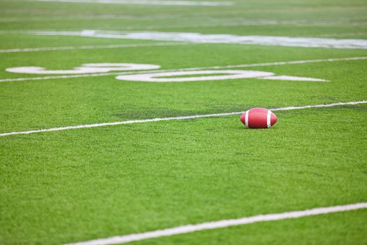 A dirty football on a professional artificial turf football field near the 30 yard line.