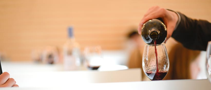 Tasting wine in a vinery-A man's hand is pouring red wine into a glass with a corkscrew set down. Lot of wine bottles in the background with shadows.