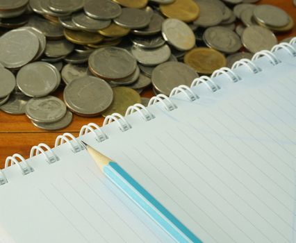 Many coins stacked on a wooden table.
And a notebook and blue pencil placed nearby.                               