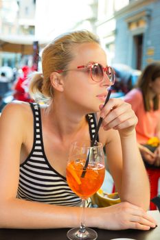 Woman terracing with aperol spritz cocktail in street cafe watching people walking by.