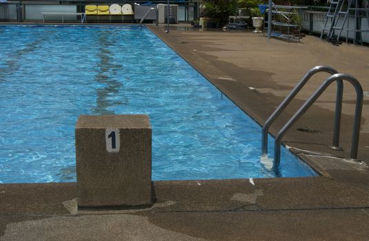 Public pool with clear blue water, stand for jump and see a ladder for access to the swimming pool.