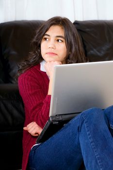 Biracial teen girl sitting against black leather couch with laptop, looking off to side,  thinking