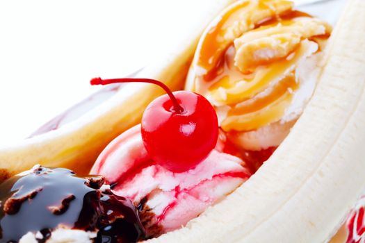 A closeup view of a delectable banana split, with chocolate, strawberry, and caramel drizzled on mounds of vanilla ice cream and topped with a red cherry.  Shot on white background.  Selective focus on cherry.