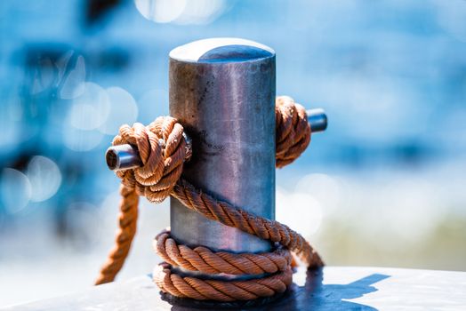 Abstract: Rope tied in a knot on a bollard. Mooring rope wrapped around the cleat on sea background. Metallic capstan in harbor for the mooring of yachts and boats. Blurred background.