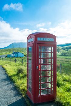 Old-fashioned traditional red telephone booth or public payphone standing next to a lonely house and a remote road in Scotland