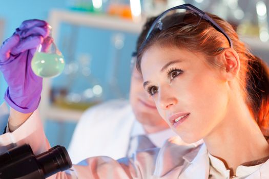 Chemical laboratory scene: attractive young student and her post doctoral supervisor scientist observing the green indikator solution color shift in glass flask.