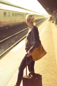 Blonde caucasian woman waiting at the railway station with a suitcase.