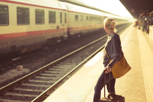 Blonde caucasian woman waiting at the railway station with a suitcase.