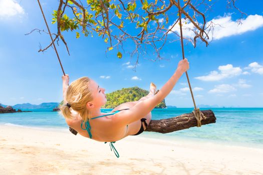 Lady swinging on the picture perfect tropical beach with the view of the island and turquoise coral reef on a sunny summer day.