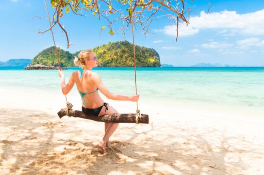 Lady swinging on the picture perfect tropical beach with the view of the island and turquoise coral reef on a sunny summer day.