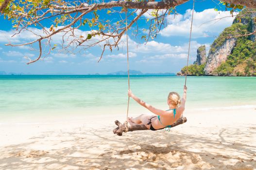 Lady swinging on the picture perfect tropical beach with the view of the island and turquoise coral reef on a sunny summer day.