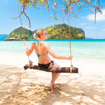 Lady swinging on the picture perfect tropical beach with the view of the island and turquoise coral reef on a sunny summer day.