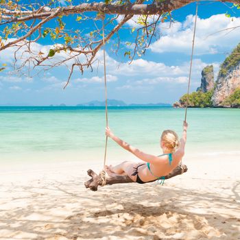 Lady swinging on the picture perfect tropical beach with the view of the island and turquoise coral reef on a sunny summer day.