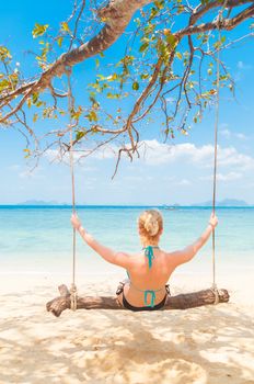 Lady swinging on the picture perfect tropical beach with the view of the island and turquoise coral reef on a sunny summer day.