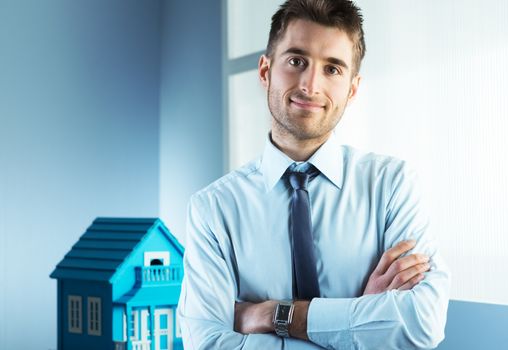 Young real estate agent with arms crossed and model house on background.