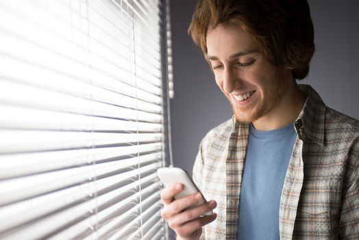 Young man smiling and using a smartphone in front of a window.