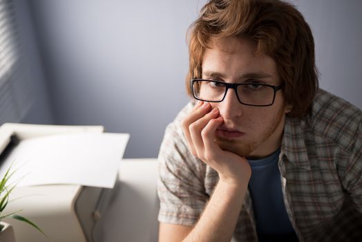 Bored young man with hand on chin and printer on background.