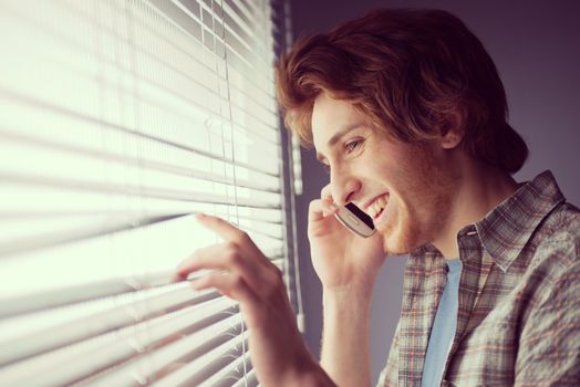 Young man smiling and talking on the phone in front of a window.