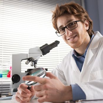 Chemical laboratory technician smiling and holding a magnifier with equipment on background.