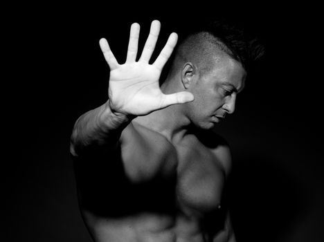 Muscular man hiding with hand raised on black background.