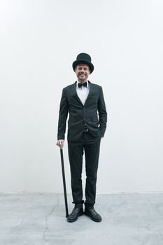 Classy smiling gentleman standing on white background and concrete floor in elegant suit with cane and bowler hat.