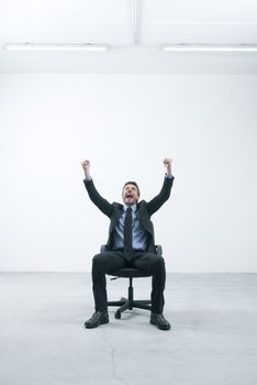 Joyful businessman with raised fists sitting on an office chair in his empty new office.