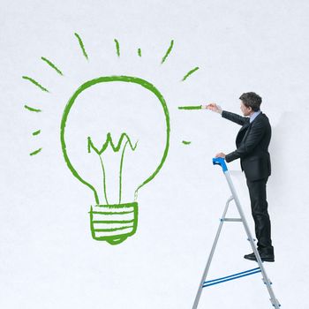 Businessman on a ladder painting a green lit bulb on a white wall.