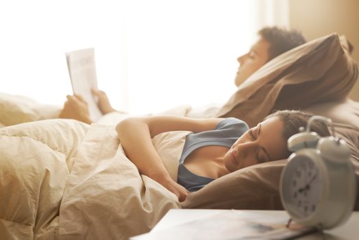 Young woman sleeping, her boyfriend is reading a newspaper on background