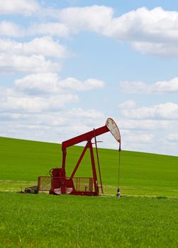 An oil well with the pump jack in action, against a grassy, green hill & cloudy blue sky.  Located in the province of Alberta, Canada.
