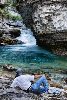 A man laying down, photographing the lower falls in Johnson Canyon, Banff National Park, Alberta, Canada, from a low angle.