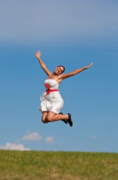 A beautiful girl in a white dress jumping for joy on a grassy, green hill, on a warm summer day.