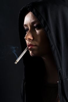 Young woman on a black background in the hood smoking a cigarette