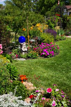 A beautiful, curving flower bed full of blooming annuals & perennials line a sunny backyard.