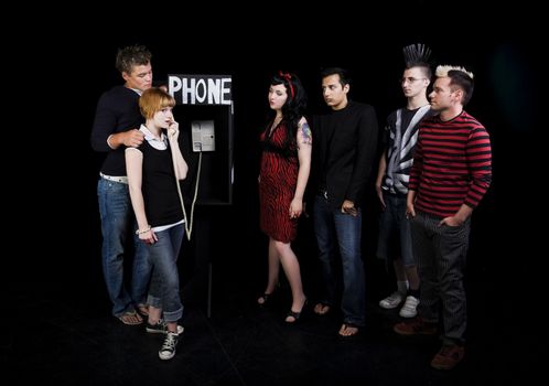 Six teenagers in a high school play.  A group of teenagers wait for their turn to use a public phone, while another girl hogs it.