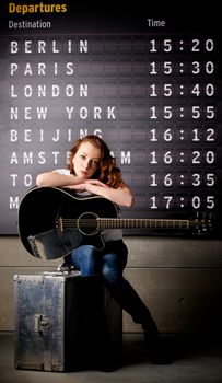 Attractive Young Woman with Guitar Sitting on Obsolete Suitcase against Arrival  Departure Board