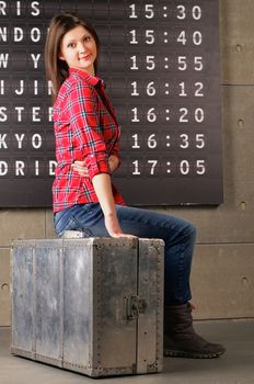 Attractive Young Woman in Checkered Shirt Sitting on Obsolete Suitcase against Arrival Departure Board