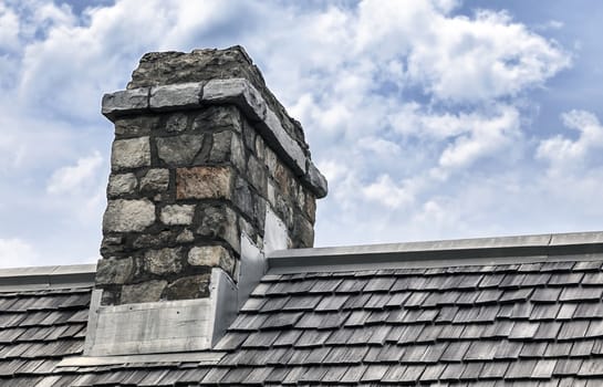 Historic chimney made of cobblestone sits on roof with wooden shingles. Beautiful cloudy background.