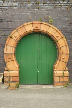 A green painted wooden door with a yellow arch designed with cast brick against an old blue brick wall with plants.