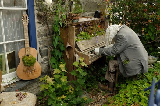 An overgrown garden with a dummy placed as a pianist with an old brocken piano and a guitar with plant growing.