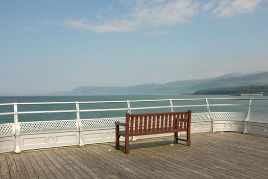 A wooden seat on boards against a silver railing looks out over the sea to mountains in the distance.