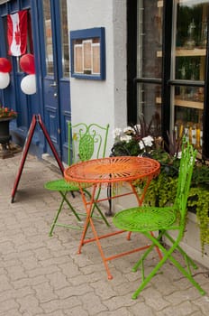 Round red metal table and a pair green metal chairs stand on the sidewalk outside the cafe