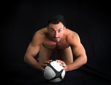 Shirtless muscular man on the floor with soccer ball, isolated on black background