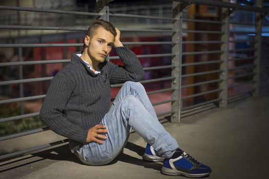 Handsome blond young man sitting alone in urban setting, looking at camera, night shot