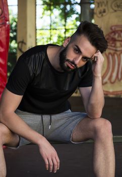 Handsome young man with beard, wearing t-shirt and shorts, looking at camera, sitting