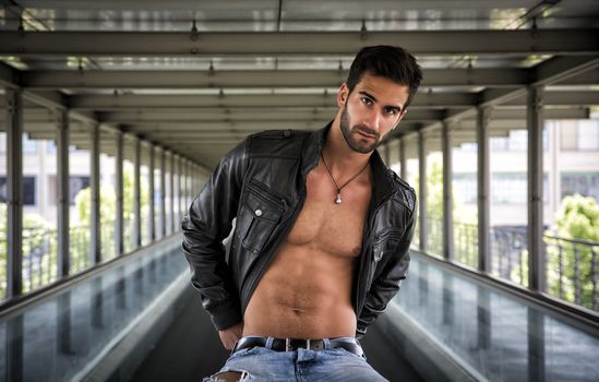 Handsome bearded young man sitting, wearing leather jacket on naked torso, outdoors in urban environment