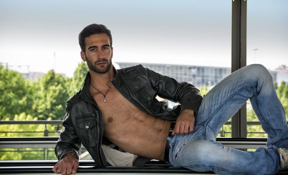 Handsome bearded young man leaning, wearing leather jacket on naked torso, outdoors in urban environment